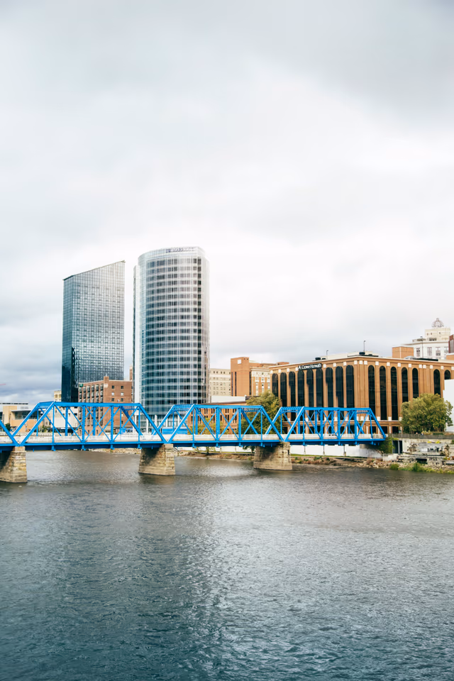 Daytime Grand Rapids skyline with the Blue Bridge and high-rises reflected in the clear blue Grand River — West Michigan Web hero background