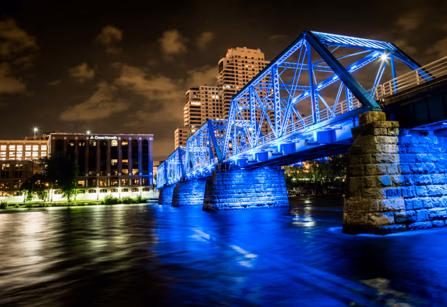 Night view from beneath the lit-up Blue Bridge over the Grand River with Grand Rapids buildings behind — West Michigan Web About hero background